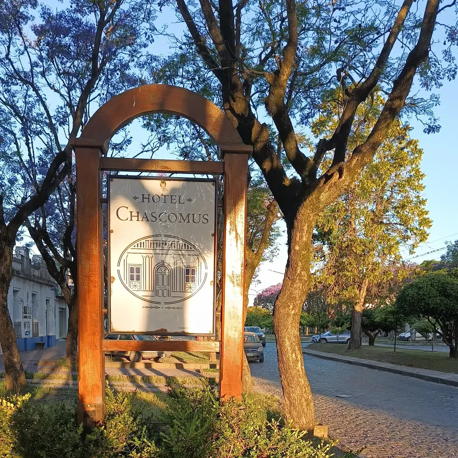El cartel del Hotel Chascomús sobre Av. Lastra, al atardecer, entre los árboles de la vereda.
