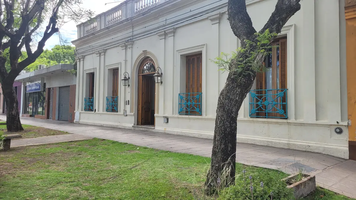 La fachada del hotel sobre Av. Lastra, con los balcones de reja turquesa y un árbol en la vereda.