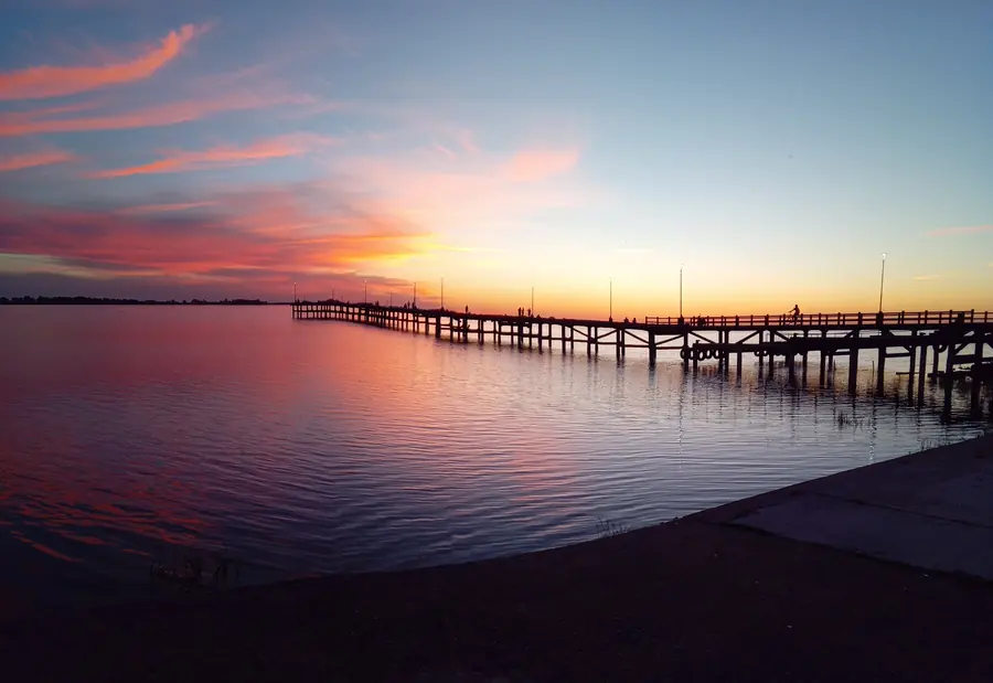 Atardecer sobre la laguna de Chascomús desde la costanera, con el cielo rosa y naranja y el muelle de pescadores entrando en el agua.